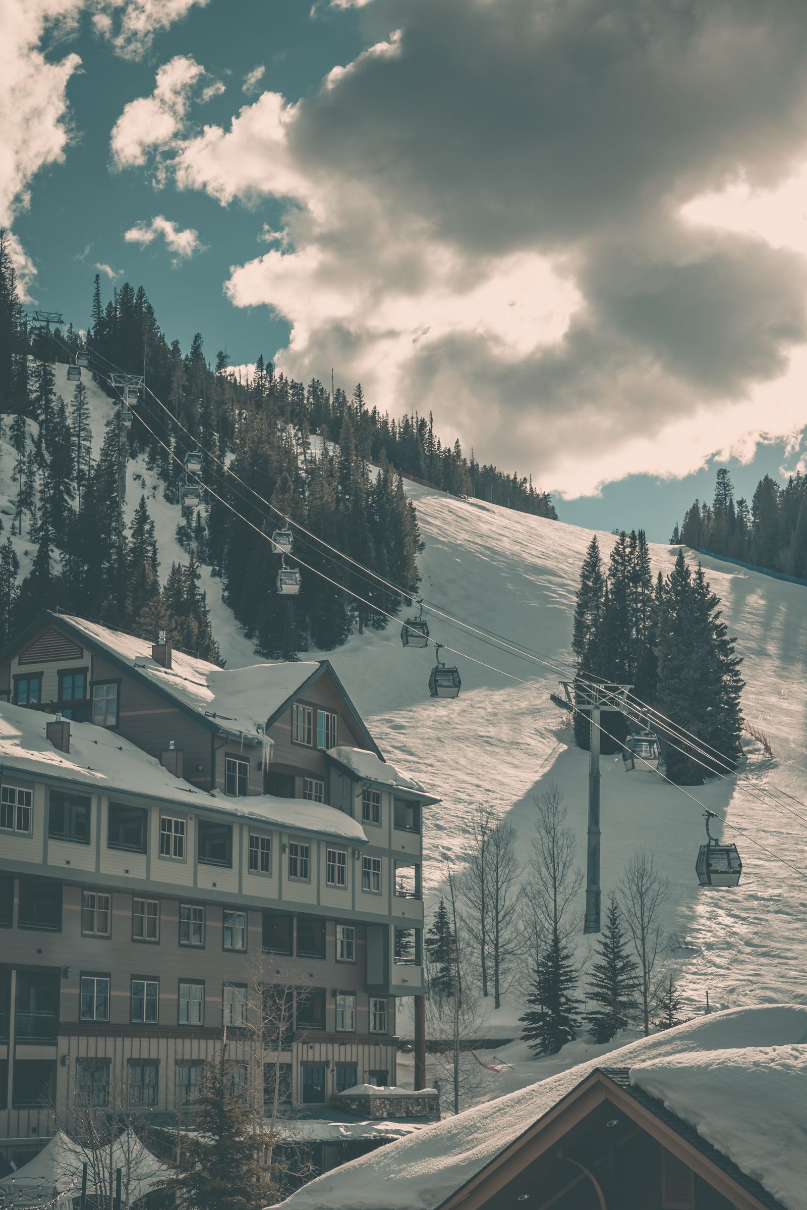 Gondola cabins climbing a snow-covered ski slope above a slopeside lodge, under a broken winter sky