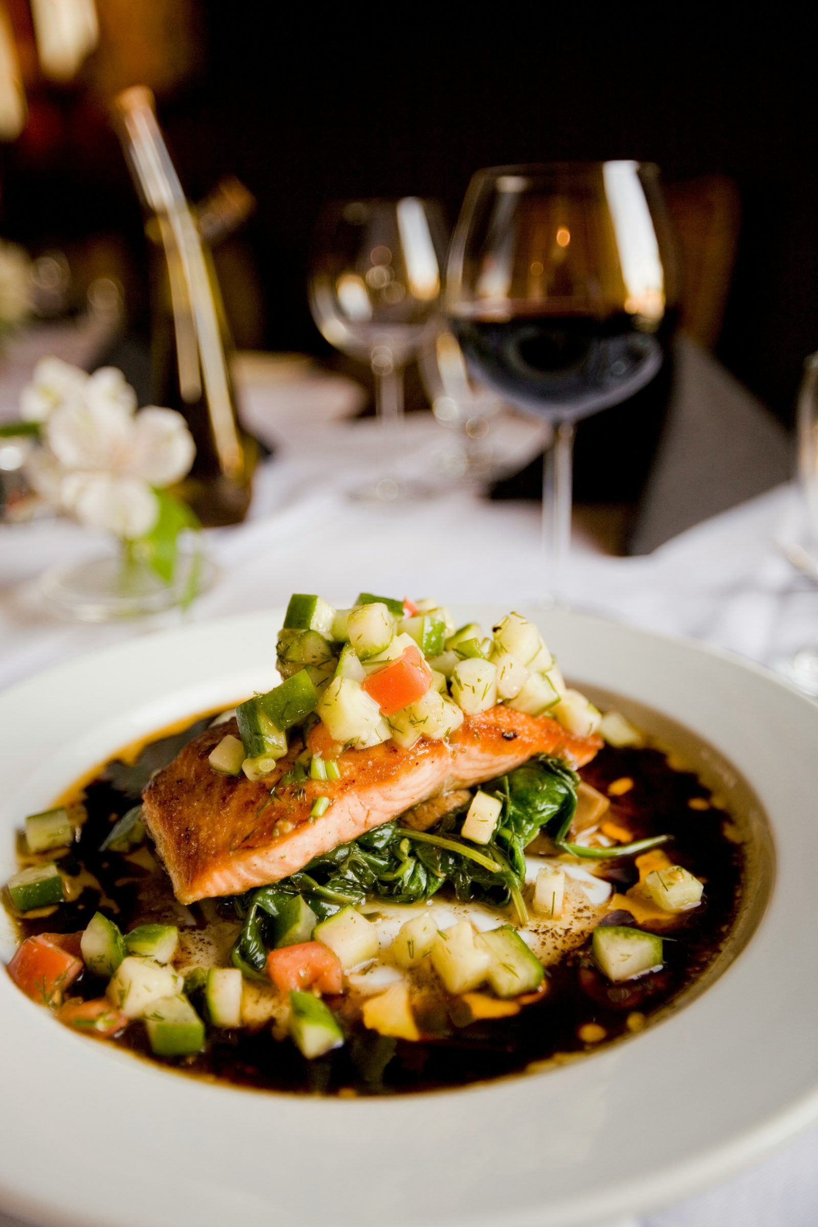 A plated fine-dining dish - seared salmon over greens with a cucumber-dill relish and a balsamic reduction, beside a glass of red wine