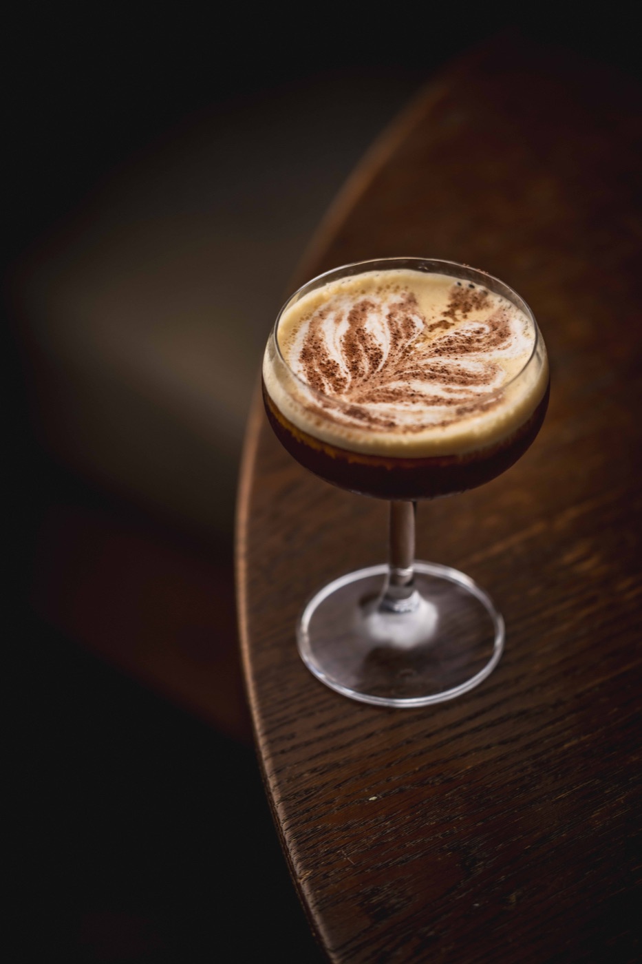 An espresso martini with a feathered foam pattern on a dark walnut table