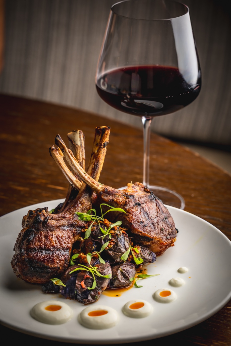 Grilled lamb chops with mushrooms and dots of sauce, beside a glass of red wine