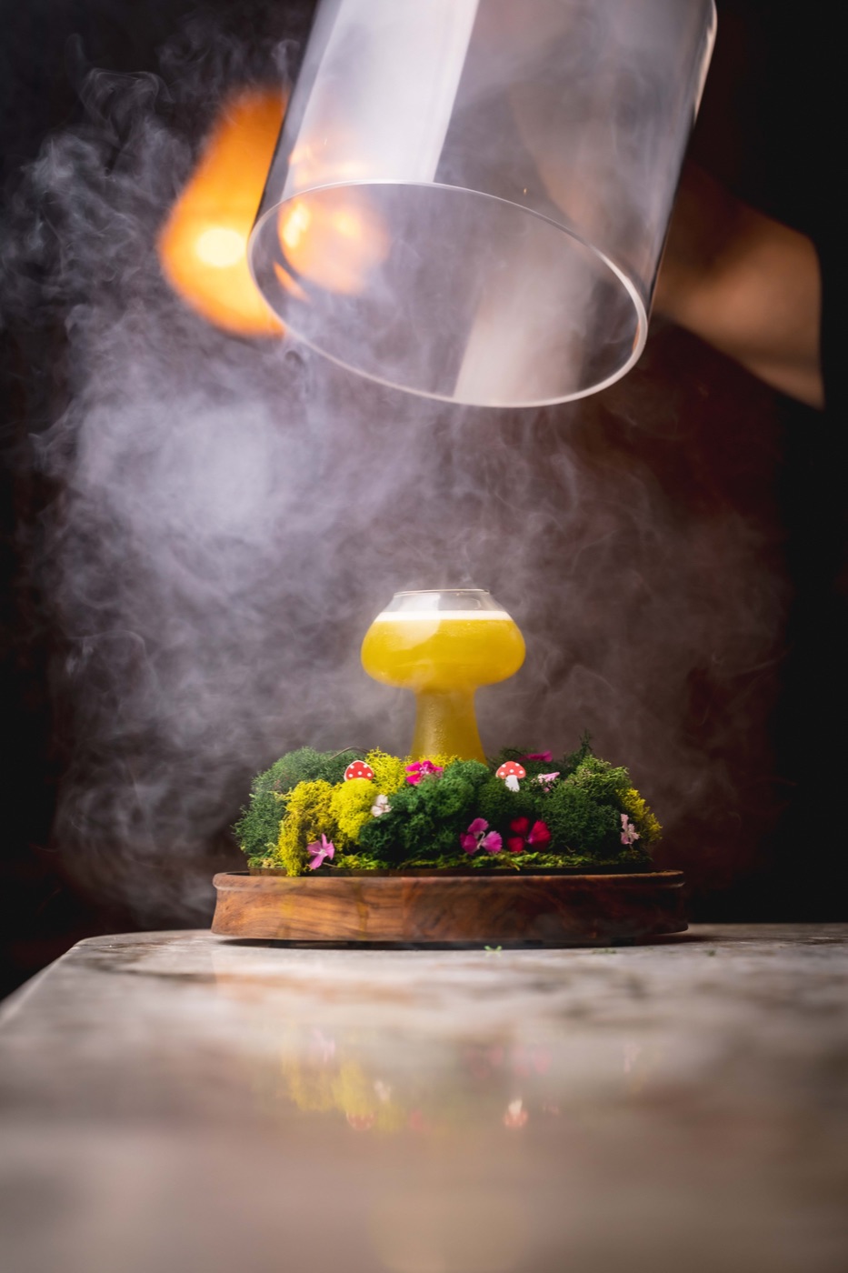 The Magic Mushroom cocktail, a yellow mushroom-shaped glass on moss and flowers, revealed from under a smoking glass cloche