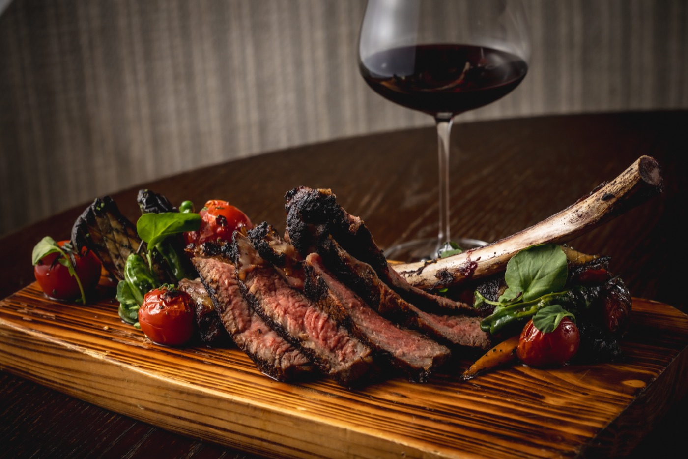 A sliced 40-day dry-aged bone-in ribeye on a wooden board with roasted tomatoes and a glass of red wine